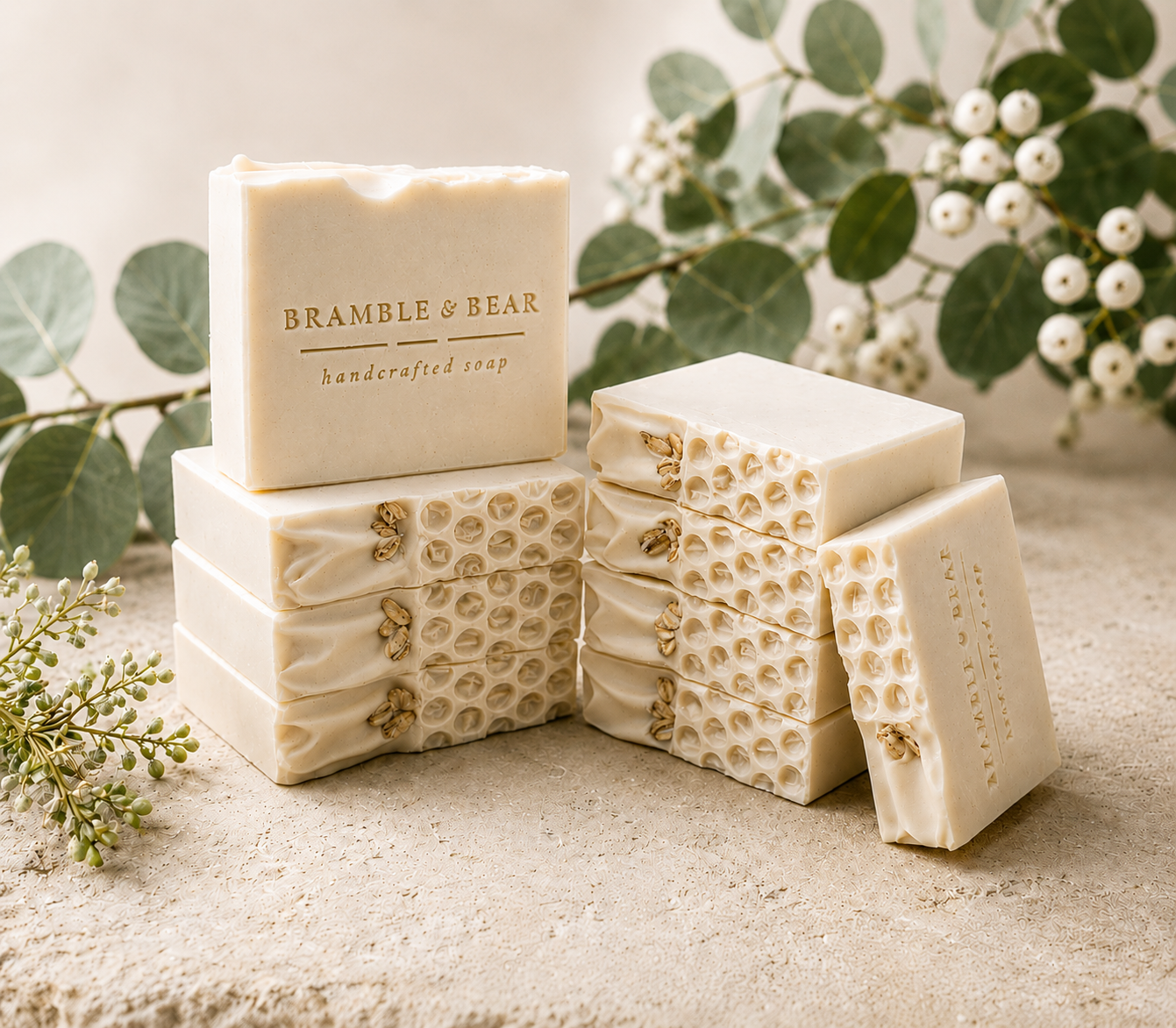 Stacked handmade Bramble and Bear Soap Co. bar soaps in soft cream with embossed logo and dotted texture, surrounded by fresh eucalyptus sprigs and white berry botanicals — Signature Soaps collection.