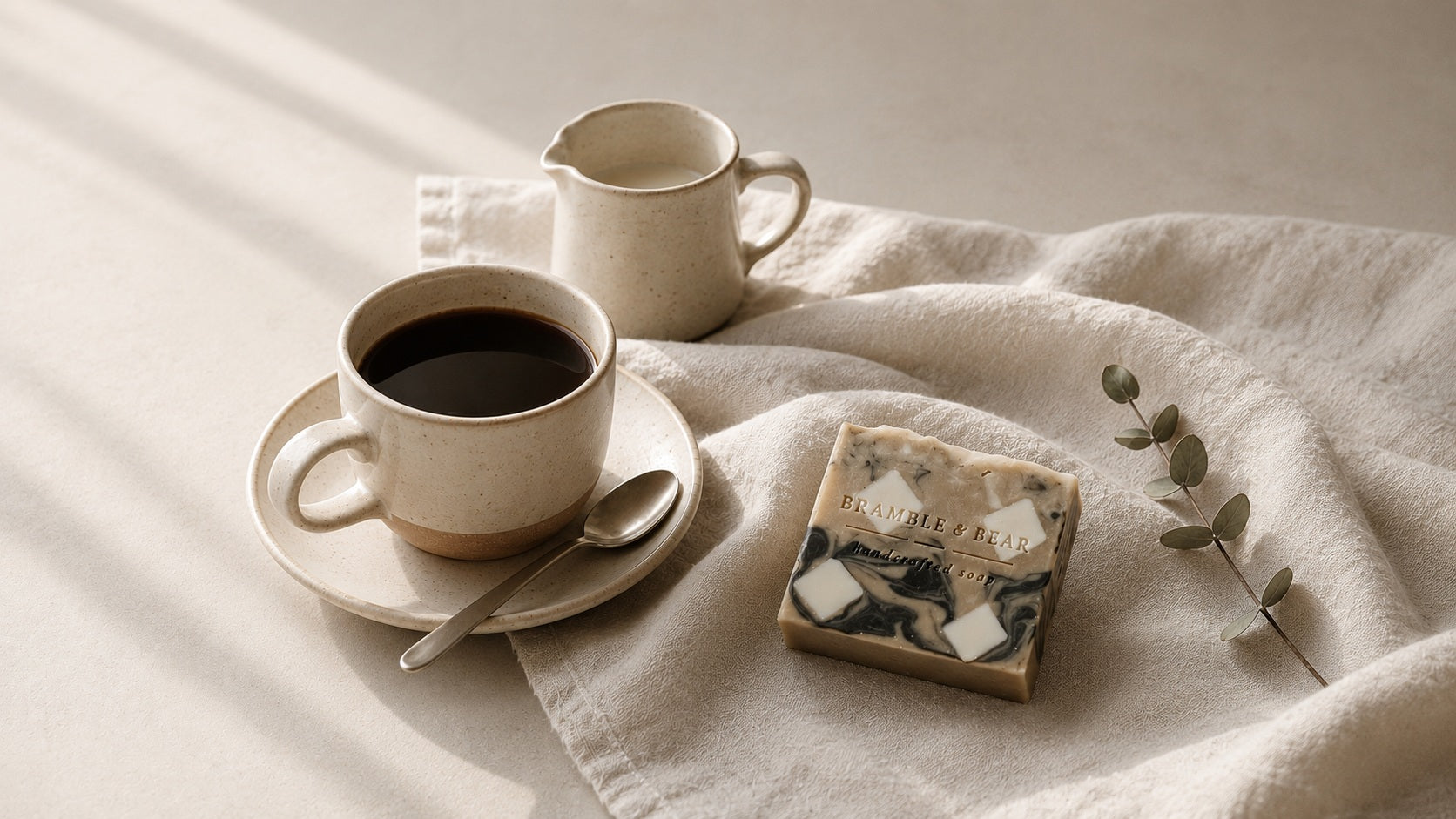 Bramble & Bear marbled soap bar beside a cream ceramic cup of coffee, saucer, spoon, and small pitcher on soft off-white linen in warm Nordic morning light.