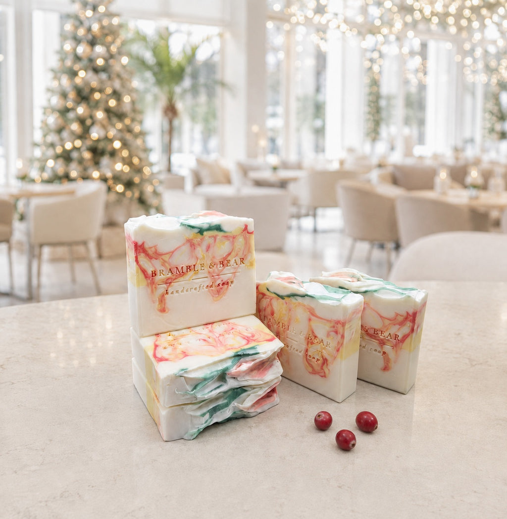 Five Bramble & Bear Cranberry Prosecco Spritz soap bars with coral, yellow, and green marbling are arranged beside three cranberries on a pale stone counter in a bright, holiday-lit hotel lounge.
