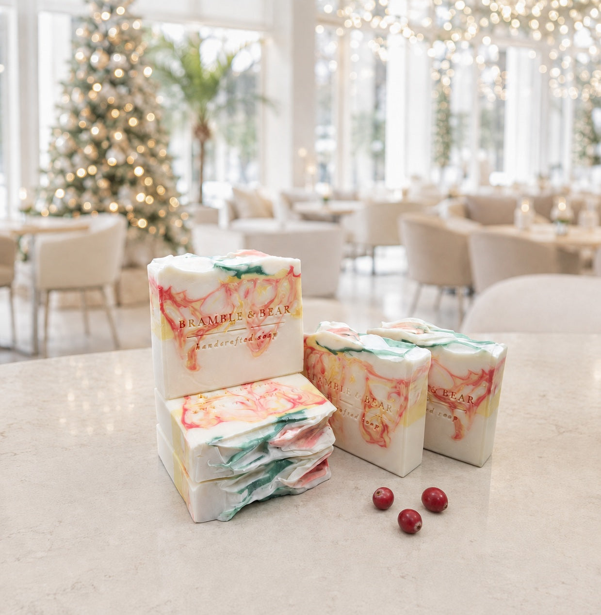Five Bramble & Bear Cranberry Prosecco Spritz soap bars with coral, yellow, and green marbling are arranged beside three cranberries on a pale stone counter in a bright, holiday-lit hotel lounge.