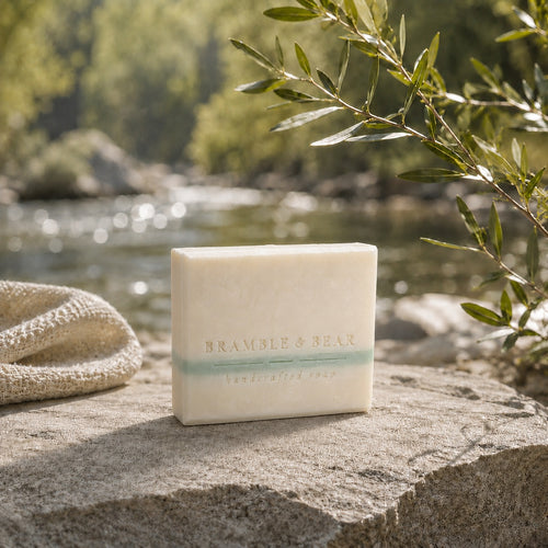 Apothecary Rice Soap on limestone beside a sunlit stream, framed by a cream cloth and leafy branch.