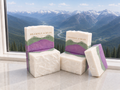Alpine Lavender Mint soap bars stacked on a marble ledge with lavender, mint green, and ivory layers, set against a bright window view of snow-capped alpine mountains and forested valleys.