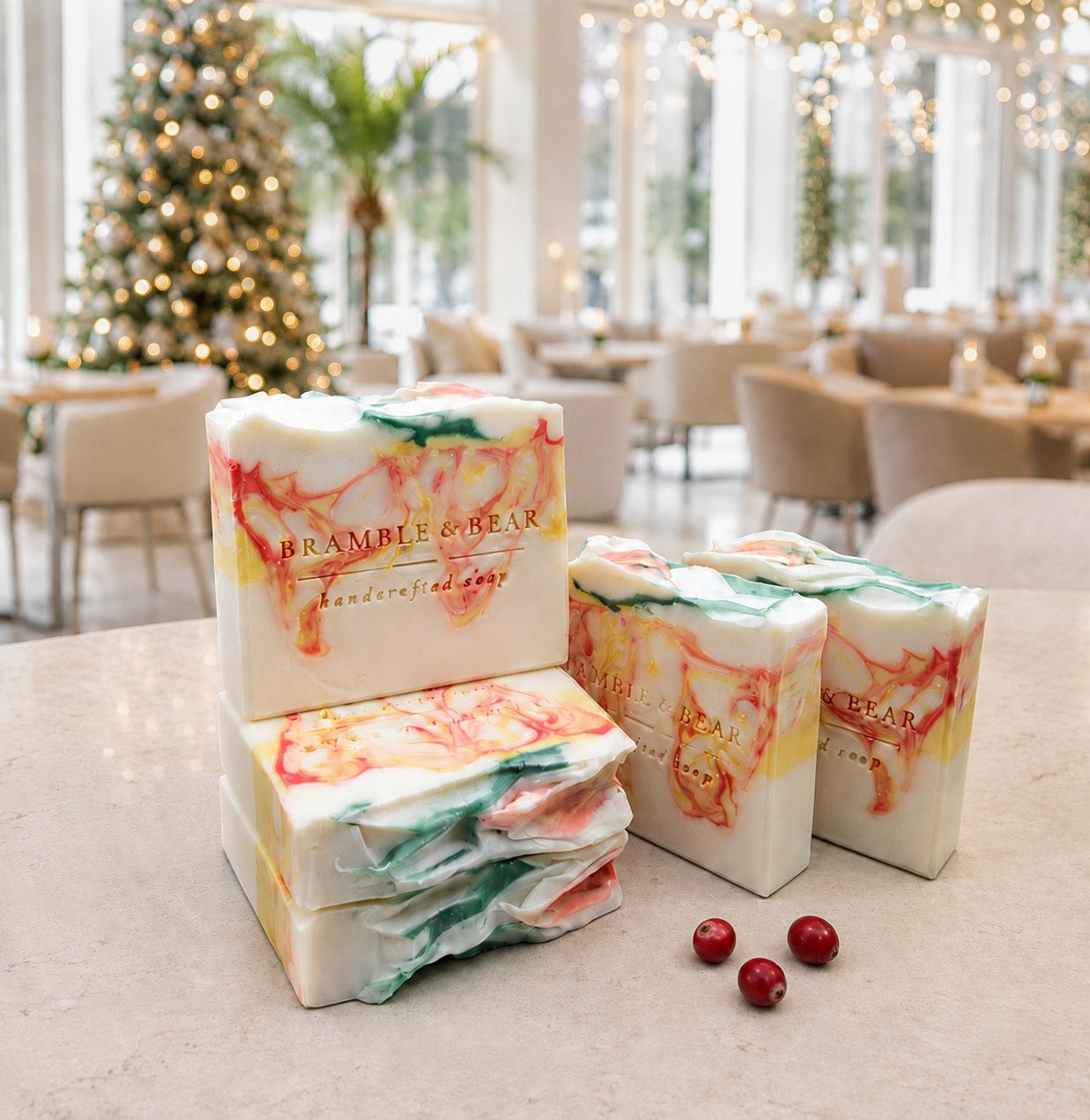 Five Bramble & Bear Cranberry Prosecco Spritz soap bars with coral, yellow, and green marbling are arranged beside three cranberries on a pale stone counter in a bright, holiday-lit hotel lounge.