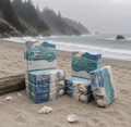 Coastal Rain Fog handmade soaps stacked on a sandy beach with blue and white ocean-inspired swirls, shell embeds, a seashell, and a misty shoreline in the background.
