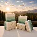 Juniper Woodland Sunset handmade soaps stacked on a marble ledge with soft green and ivory layers, whipped mountain-like tops, evergreen forest, alpine peaks, and warm sunset light.