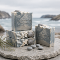 Northern Fjord Air handmade soaps arranged on a stone surface near the coast, with blue-gray, cream, and charcoal swirls, gray pebble embeds, small stones, driftwood, and a misty ocean background.