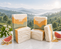 Sunlit Citrus Grove soap bars stacked on a marble ledge with ivory, apricot, and sage green layers, styled with citrus blossoms, grapefruit, and soft rolling hills in the background.