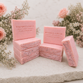 Stacked Wild Rose Meadow soap bars in soft pink tones, arranged with roses and delicate meadow flowers on a light stone surface.