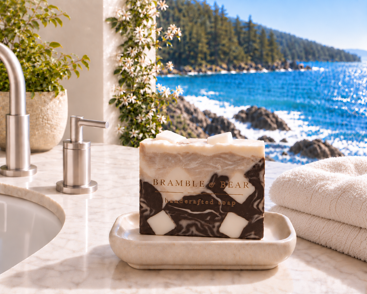 Single Bramble & Bear Iced Caramel Macchiato soap bar on a cream ceramic dish by a marble sink, with folded towels and a sunlit ocean view behind it.
