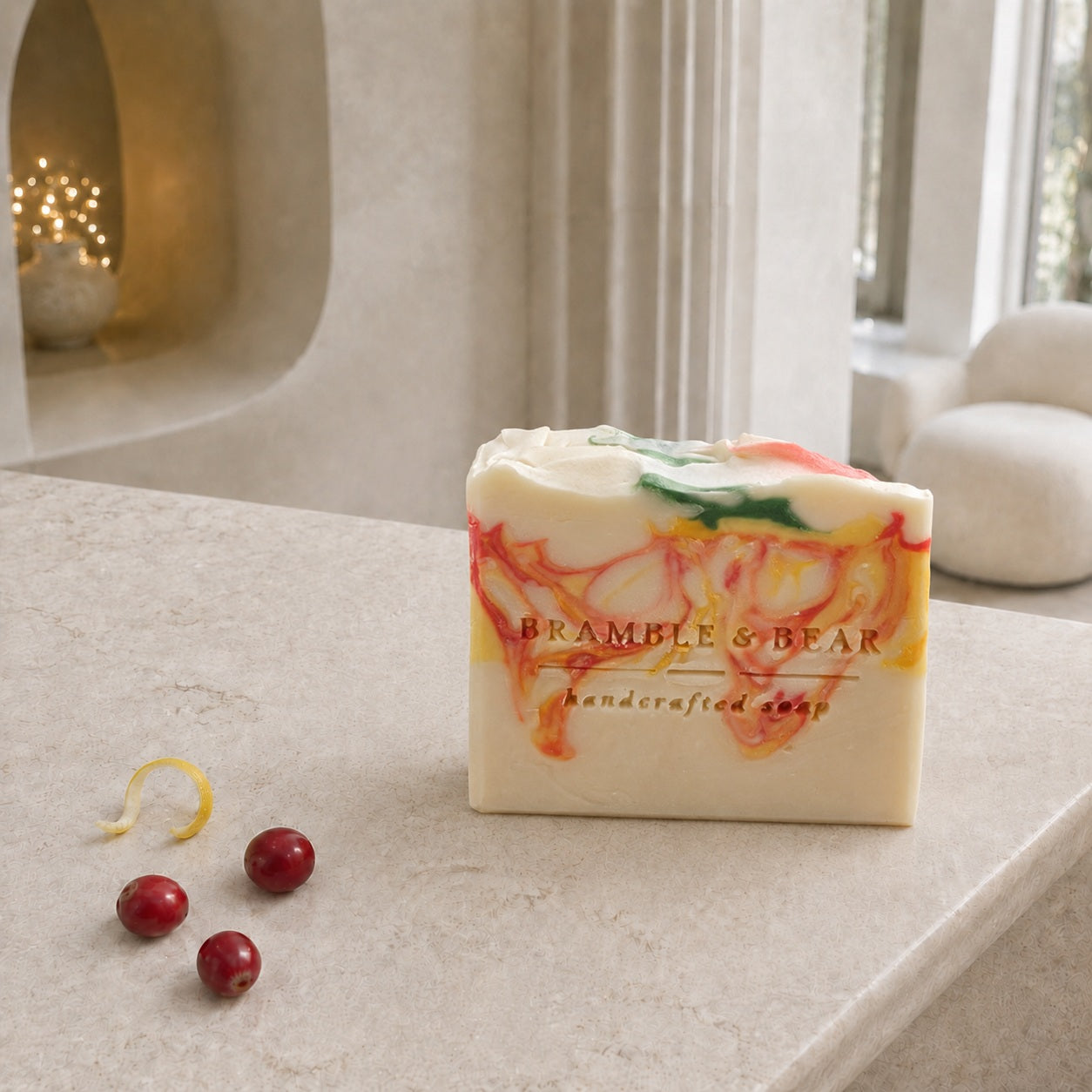 A Bramble & Bear Cranberry Prosecco Spritz soap bar with coral, yellow, and green marbling sits beside three cranberries and a curled citrus rind on a pale stone counter in an elegant ivory hotel lounge.