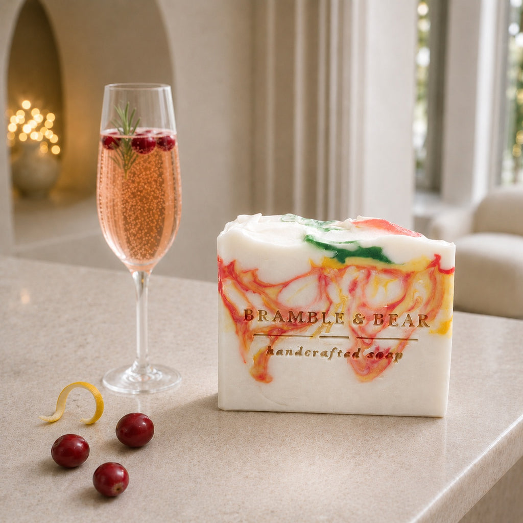 A Bramble & Bear Cranberry Prosecco Spritz soap bar with coral, yellow, and green marbling sits beside three cranberries and a curled citrus rind on a pale stone counter in an elegant ivory hotel lounge.