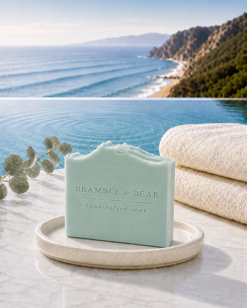 Coastal Eucalyptus Mist handmade soap standing on a round dish beside eucalyptus and folded towels, with a soft aqua color and an infinity pool overlooking a blurred coastline.