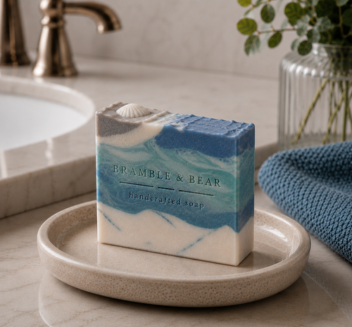 Coastal Rain Fog handmade soap resting on a round dish in a bright bathroom setting, with blue and white wave-like swirls, a shell embed, a towel, and a blurred sink faucet nearby.