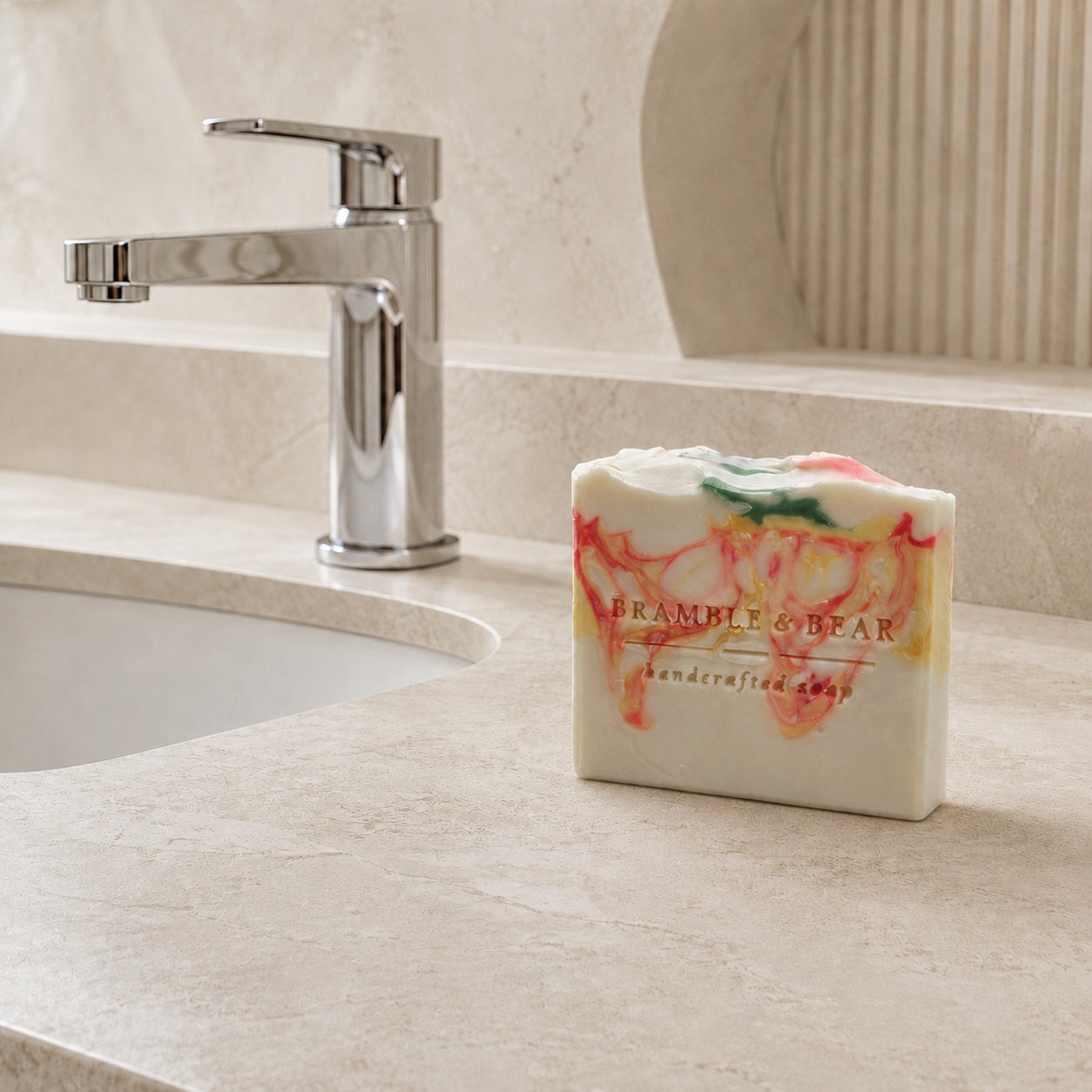 A Bramble & Bear Cranberry Prosecco Spritz soap bar with coral, yellow, and green marbling sits beside a chrome faucet on a pale marble counter in a contemporary hotel bathroom.
