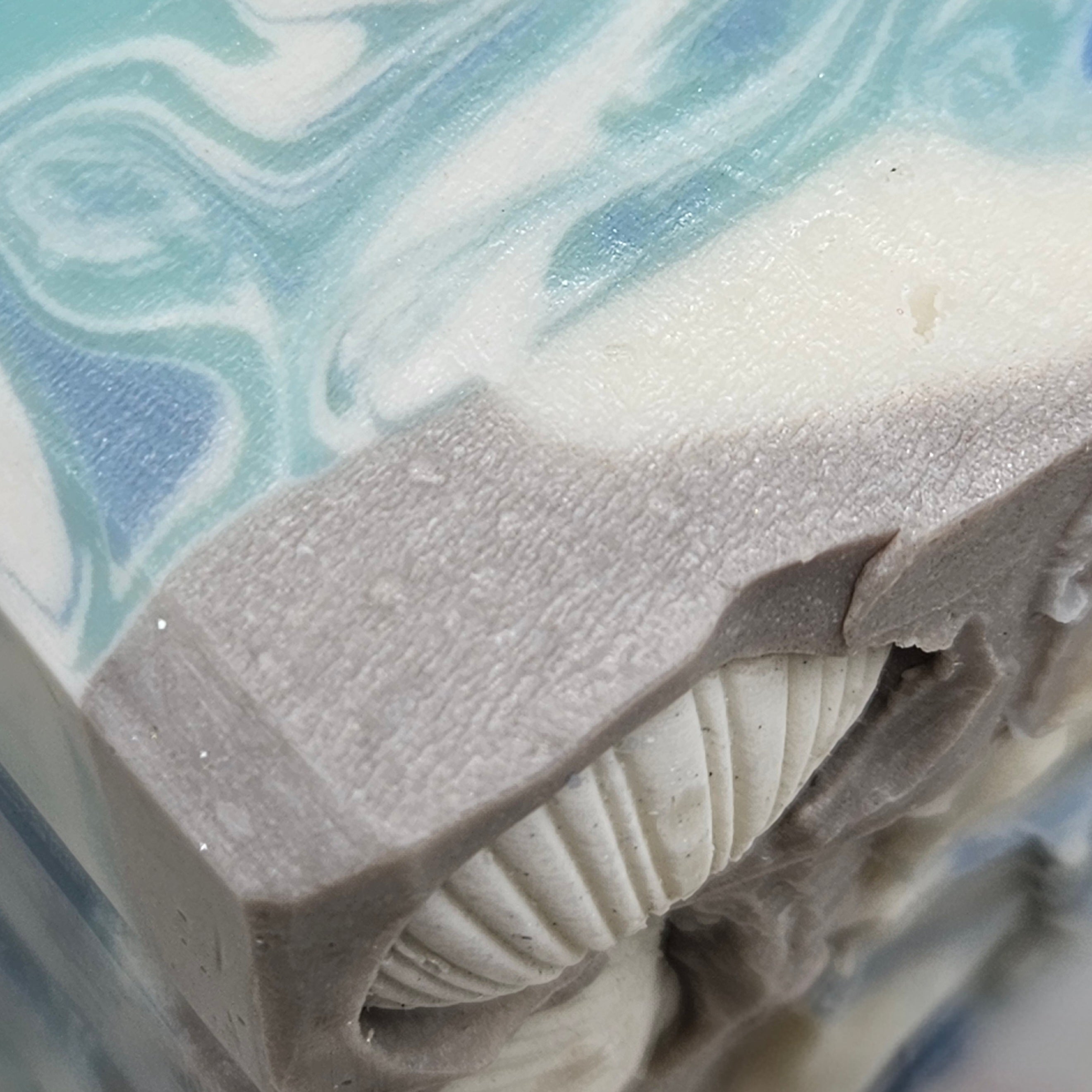 Close-up of Coastal Rain Fog handmade soap showing blue and white wave-like swirls, a gray textured layer, and a shell embed detail.