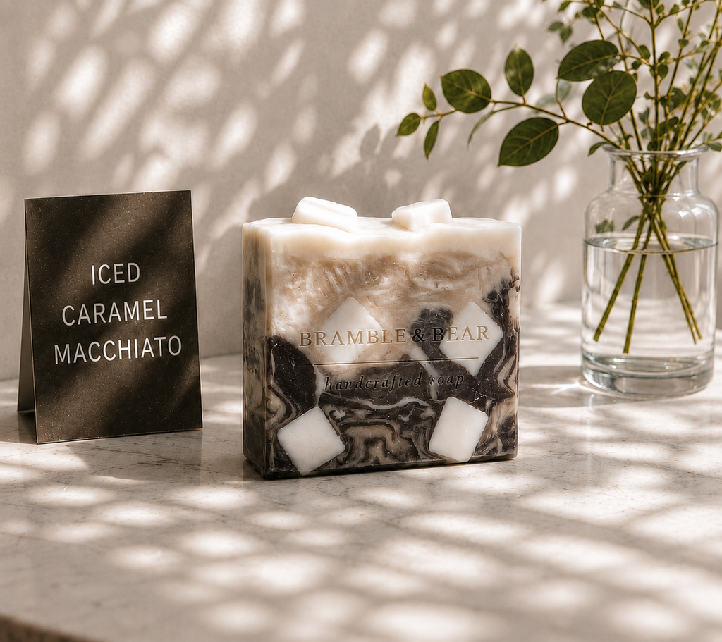 Bramble & Bear Iced Caramel Macchiato soap displayed on a marble surface beside a black product card and glass vase, with soft leaf shadows across the scene.