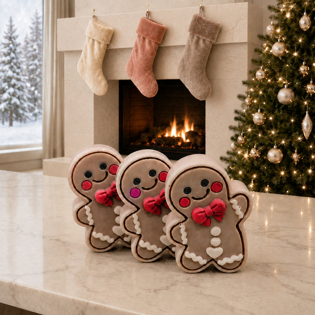 Three Frosted Gingerbread Cutie soaps on a pale stone counter before a fireplace with three stockings, a lit Christmas tree and snowy window.