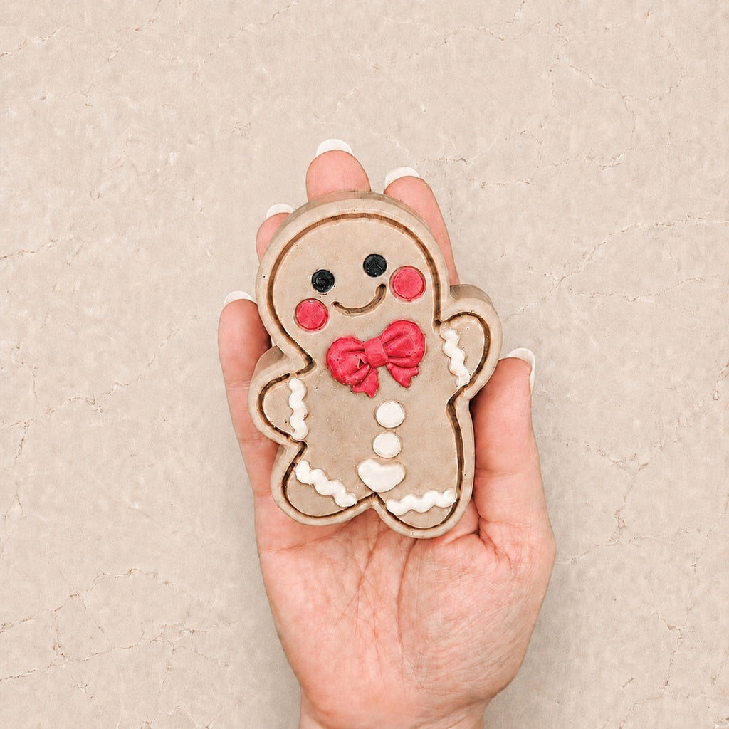 A hand holding a Frosted Gingerbread Cutie with a beige body, black eyes, red cheeks and bow, and white icing-style details, against a textured beige background.