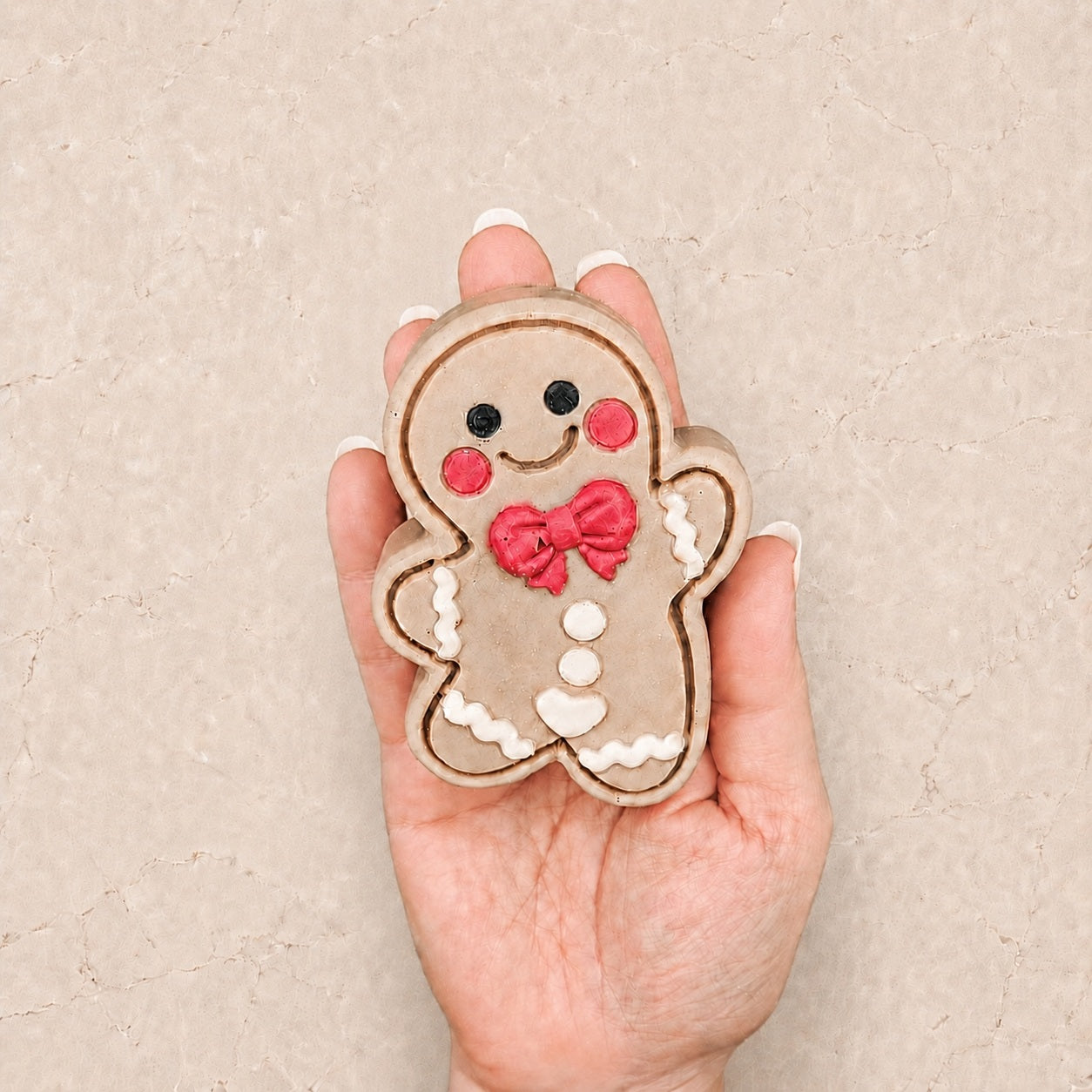 A hand holding a Frosted Gingerbread Cutie with a beige body, black eyes, red cheeks and bow, and white icing-style details, against a textured beige background.