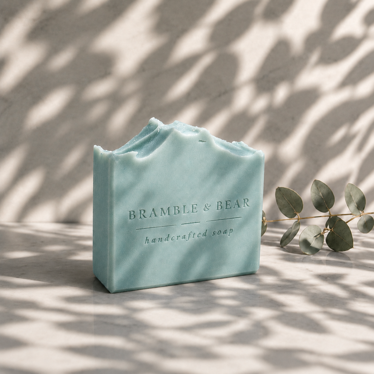 Coastal Eucalyptus Mist handmade soap on a marble surface with soft aqua color, eucalyptus, recessed stamp details, and dappled botanical shadows across the scene.
