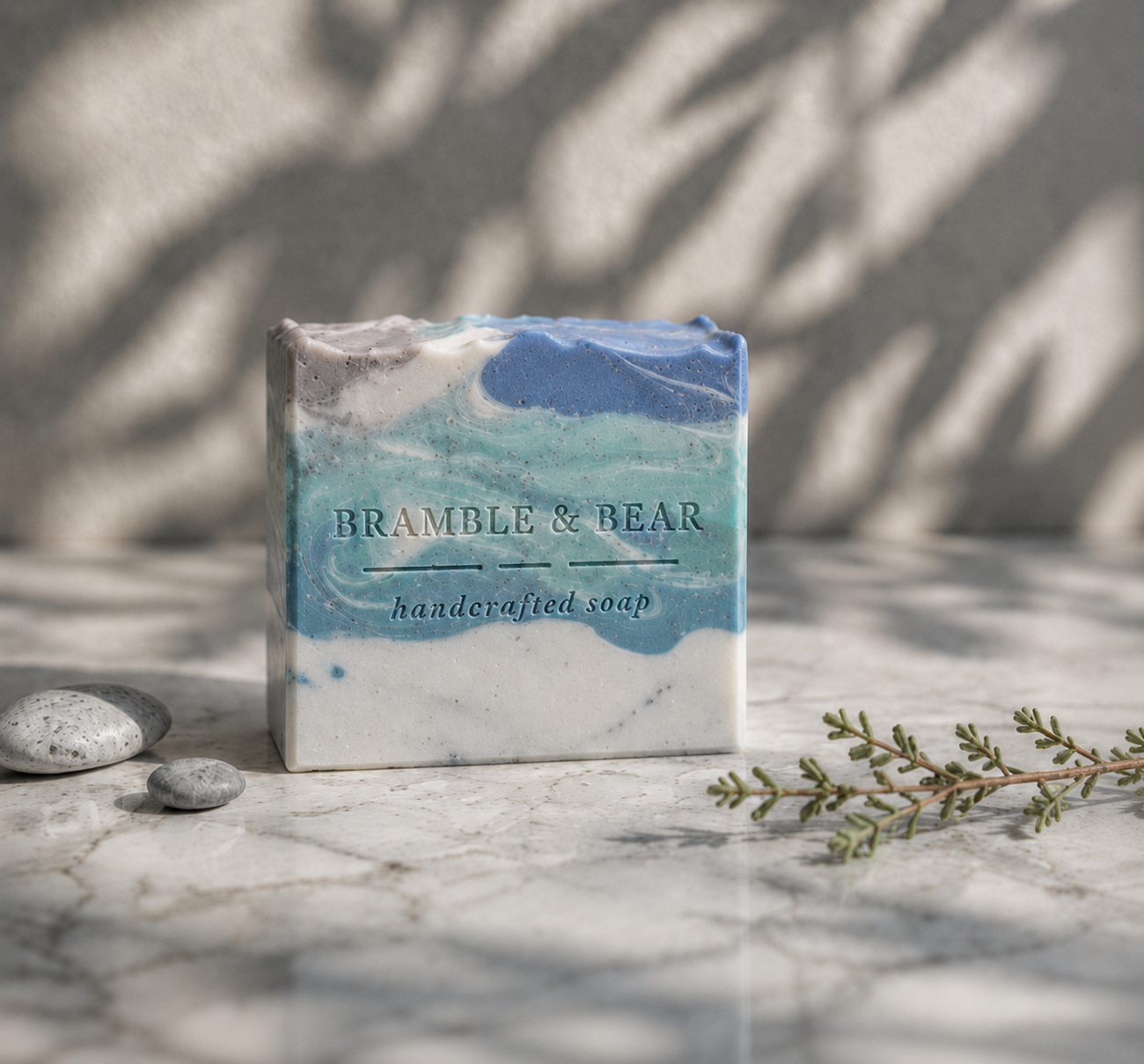 Coastal Rain Fog handmade soap on a bright surface with blue and white coastal swirls, a shell embed, small seashells, soft greenery, and botanical shadows on the wall.