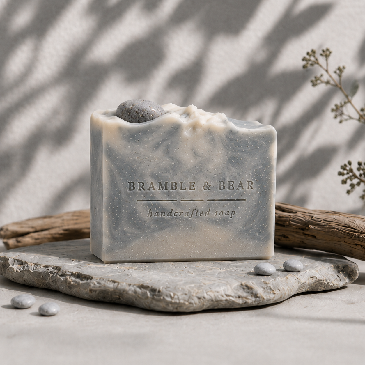 Northern Fjord Air handmade soap on a light stone surface with blue-gray and cream swirls, a gray pebble embed, driftwood, small stones, delicate greenery, and soft botanical shadows on the wall.