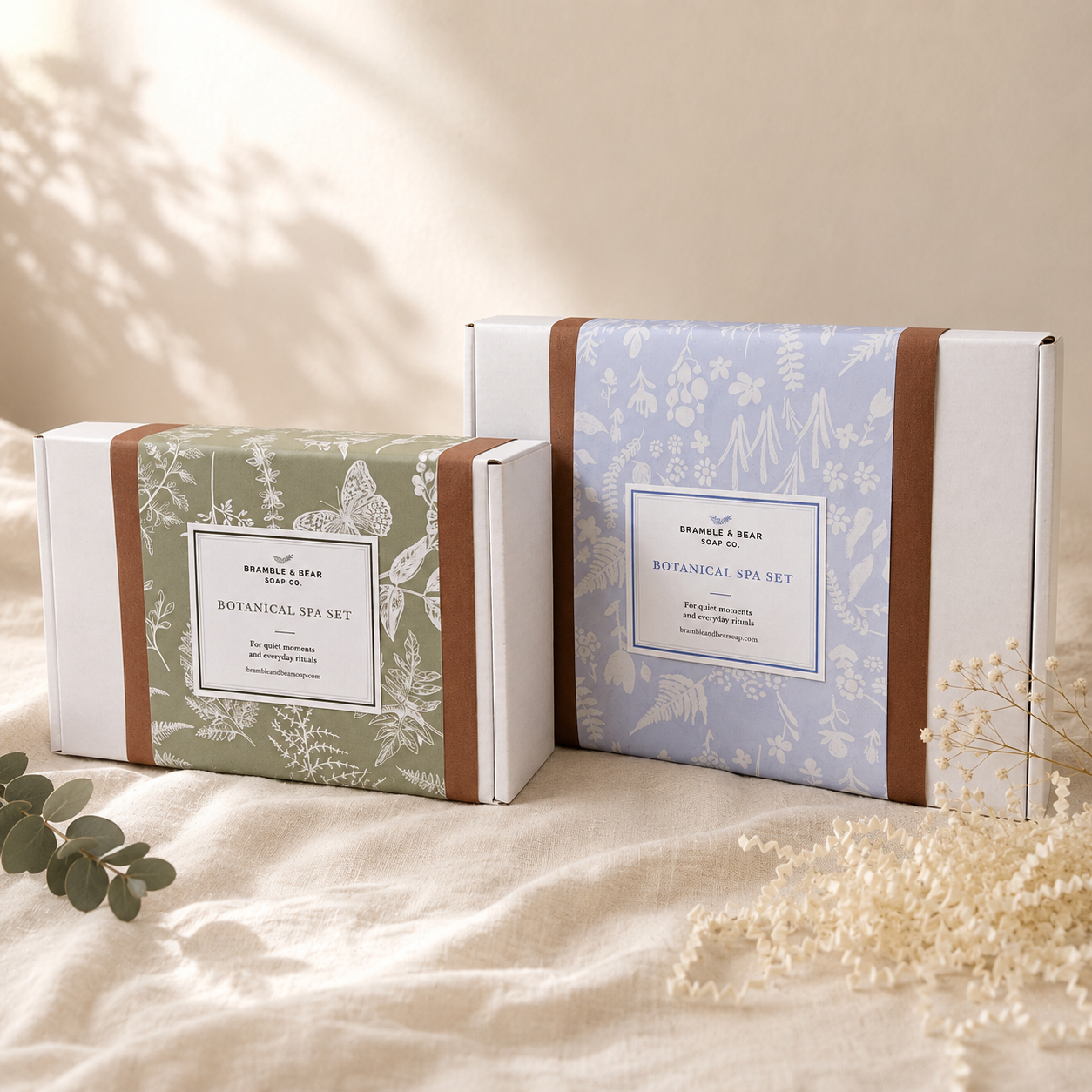 Small sage and larger pale blue Bramble & Bear botanical gift boxes displayed together on warm ivory linen with eucalyptus and dried flowers.