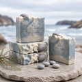 Northern Fjord Air handmade soaps arranged on a stone surface near the coast, with blue-gray, cream, and charcoal swirls, gray pebble embeds, small stones, driftwood, and a misty ocean background.