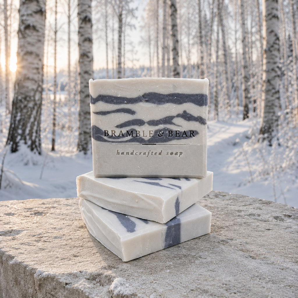 Silver Birch Crossing soap bars stacked on pale stone before a softly blurred, snow-covered birch forest.